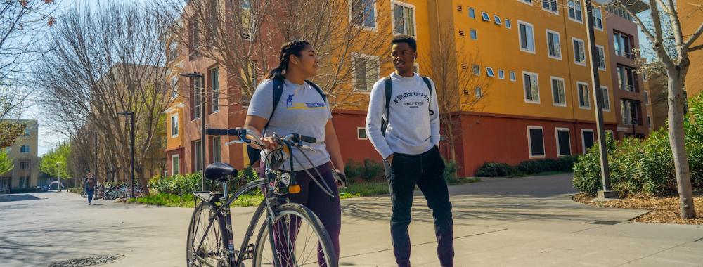 Two students walk outside the Tercero residence halls. One students walks their bike next to them.